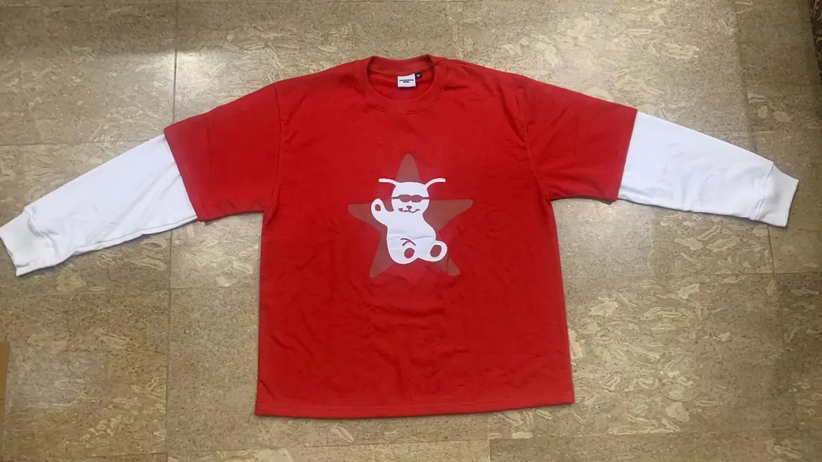 A finished red long-sleeve tee with white layered sleeves and a printed graphic, laid flat on a tiled floor